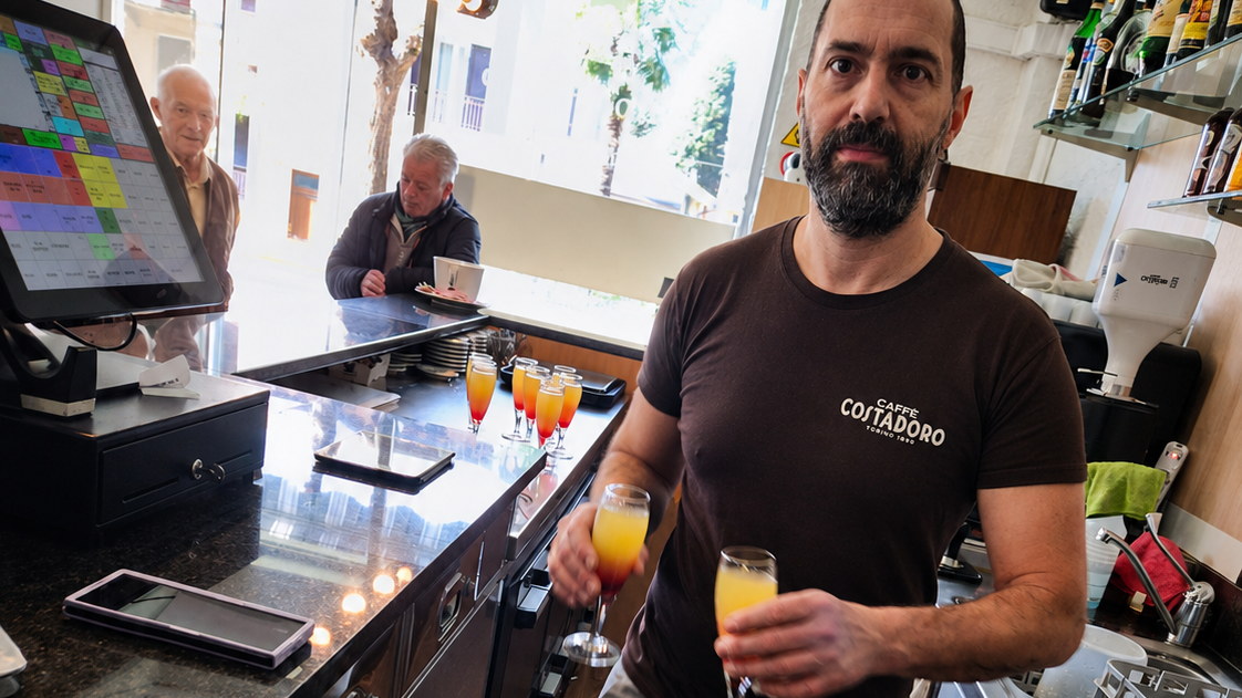 Leonardo dietro il banco del bar con due calici di aperitivo, clienti al bancone